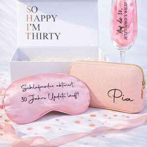 Personalisierte Geschenkbox zum 30. Geburtstag für Frauen mit rosa Kosmetiktasche mit Name Pia, Schlafmaske mit Aufdruck „Schlafmodus aktiviert, 30 Jahre Update läuft“ und Sektglas mit Spruch „Auf die 30, jetzt wird’s wild“, dekoriert mit Konfetti und Geschenkbox im Hintergrund