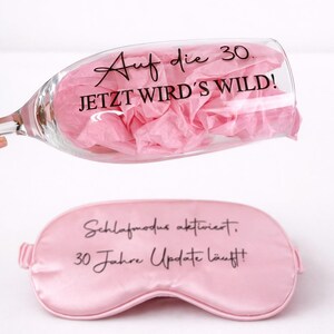 Sektglas mit Aufdruck ‚Auf die 30 – jetzt wird’s wild!‘ und rosa Schlafmaske als Geschenkset zum 30. Geburtstag vor hellem Hintergrund