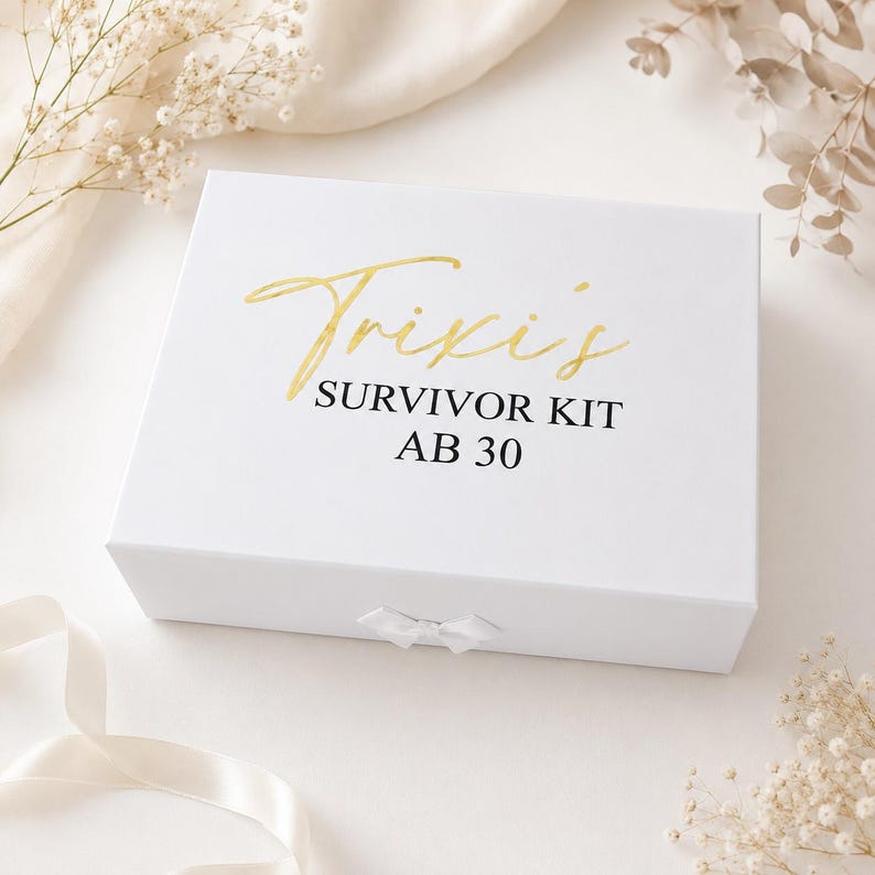 Weiße Geschenkbox mit goldener Aufschrift „Trixis Survivor Kit ab 30“ auf hellem, neutralem Hintergrund mit dezenter Deko aus Schleifenband und Trockenblumen.
