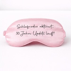Rosa Schlafmaske mit Aufdruck ‚Schlafmodus aktiviert, 30 Jahre Update läuft!‘ vor hellem, minimalistischem Hintergrund
