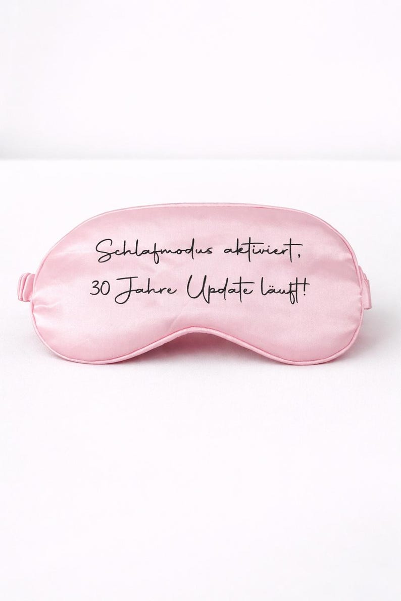 Rosa Schlafmaske mit Aufdruck ‚Schlafmodus aktiviert, 30 Jahre Update läuft!‘ vor hellem, minimalistischem Hintergrund