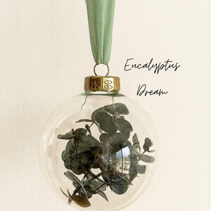 Personalisierte Weihnachtskugel aus Glas mit goldenem Kranz und grünen Chiffon-Aufhänger, gefüllt mit Eukalyptus-Trockenblättern, Variante ‚Eucalyptus Dream‘.