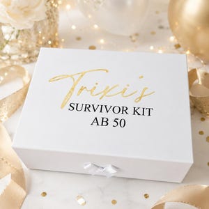 Weiße Geschenkbox mit goldener Aufschrift „Trixis Survivor Kit ab 50“ in feierlicher Dekoration mit goldenen Bändern, Konfetti, Blumen und Ballons auf hellem Hintergrund.