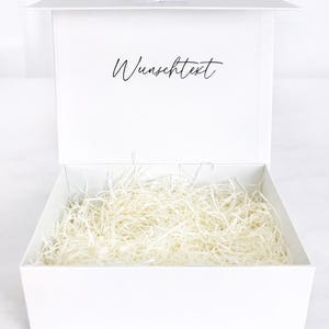 Weiße offene Geschenkbox mit neutralem Füllmaterial aus weißer Holzwolle, Innenseite mit Schriftzug „Wunschtext“, auf hellem Hintergrund.