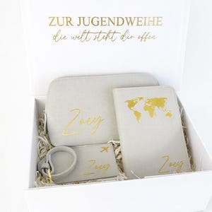 Personalisierte Geschenkbox zur Jugendweihe mit grauem Reiseset bestehend aus Kosmetiktasche, Reisepasshülle und Kofferanhänger in Gold mit Namen Zoey auf hellem, neutralem Hintergrund