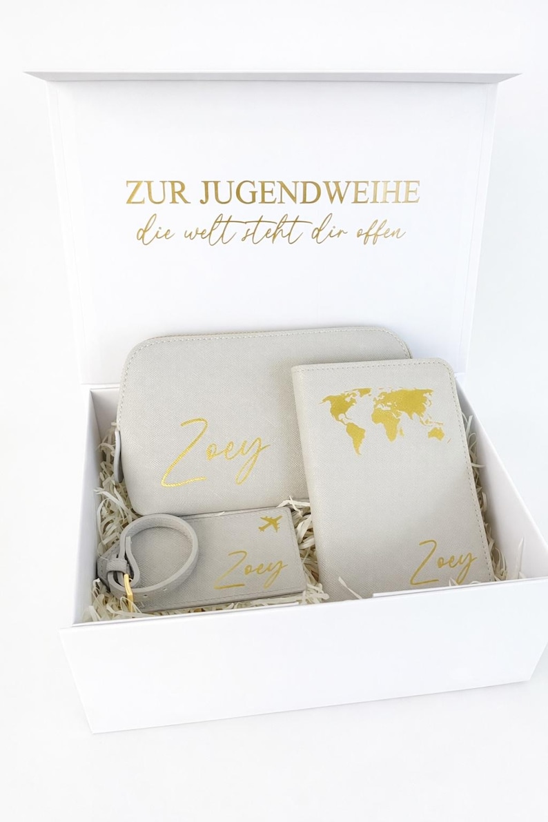 Personalisierte Geschenkbox zur Jugendweihe mit grauem Reiseset bestehend aus Kosmetiktasche, Reisepasshülle und Kofferanhänger in Gold mit Namen Zoey auf hellem, neutralem Hintergrund