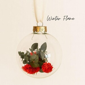 Personalisierte Weihnachtskugel aus Glas mit goldenem Kranz und weißen Chiffon-Aufhänger, gefüllt mit roten Phalaris-Trockenblumen und grünem Eukalyptus, Variante ‚Winter Flame‘.