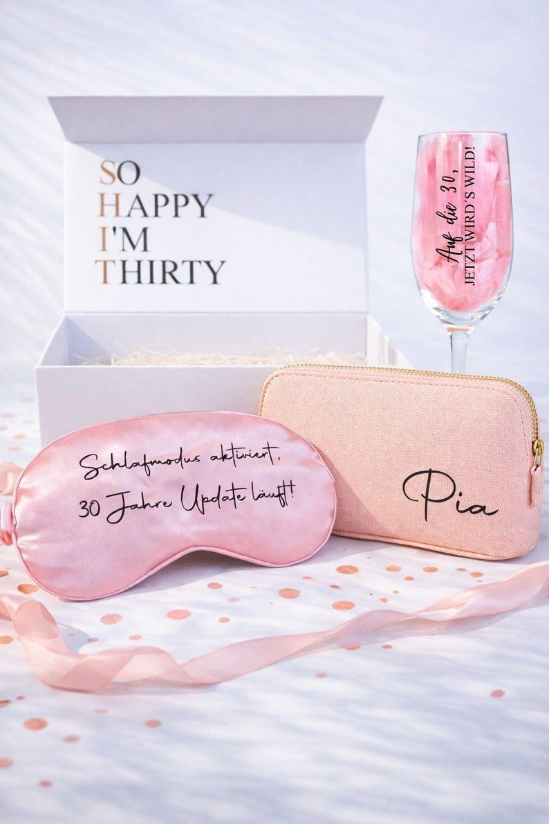 Personalisierte Geschenkbox zum 30. Geburtstag für Frauen mit rosa Kosmetiktasche mit Name Pia, Schlafmaske mit Aufdruck „Schlafmodus aktiviert, 30 Jahre Update läuft“ und Sektglas mit Spruch „Auf die 30, jetzt wird’s wild“, dekoriert mit Konfetti und Geschenkbox im Hintergrund