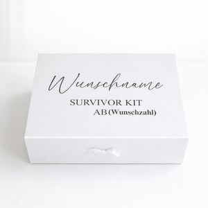 Weiße personalisierte Geschenkbox ‚Survivor Kit ab (Wunschzahl)‘ mit Schleife auf hellem, minimalistischem Hintergrund