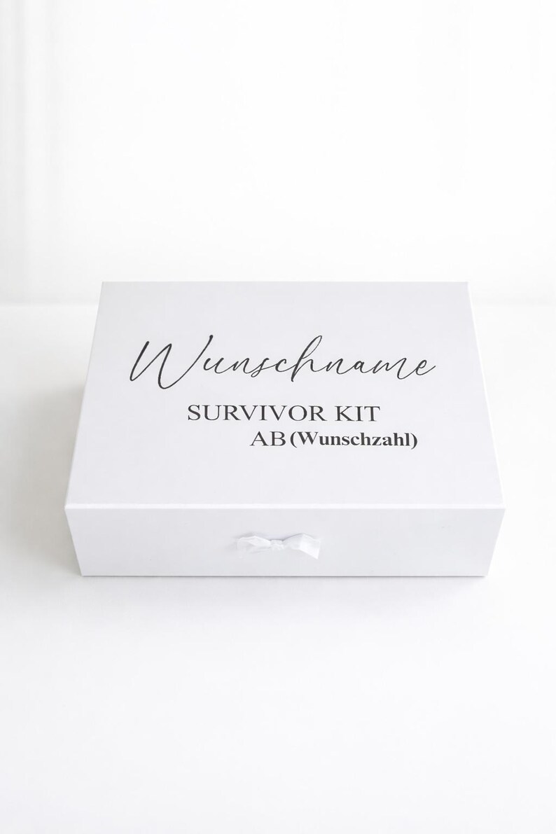 Weiße personalisierte Geschenkbox ‚Survivor Kit ab (Wunschzahl)‘ mit Schleife auf hellem, minimalistischem Hintergrund