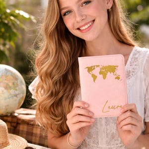 Glückliches Mädchen hält eine personalisierte rosa Reisepasshülle mit Weltkartenmotiv in den Händen vor modernem Reisehintergrund mit Globus und Accessoires