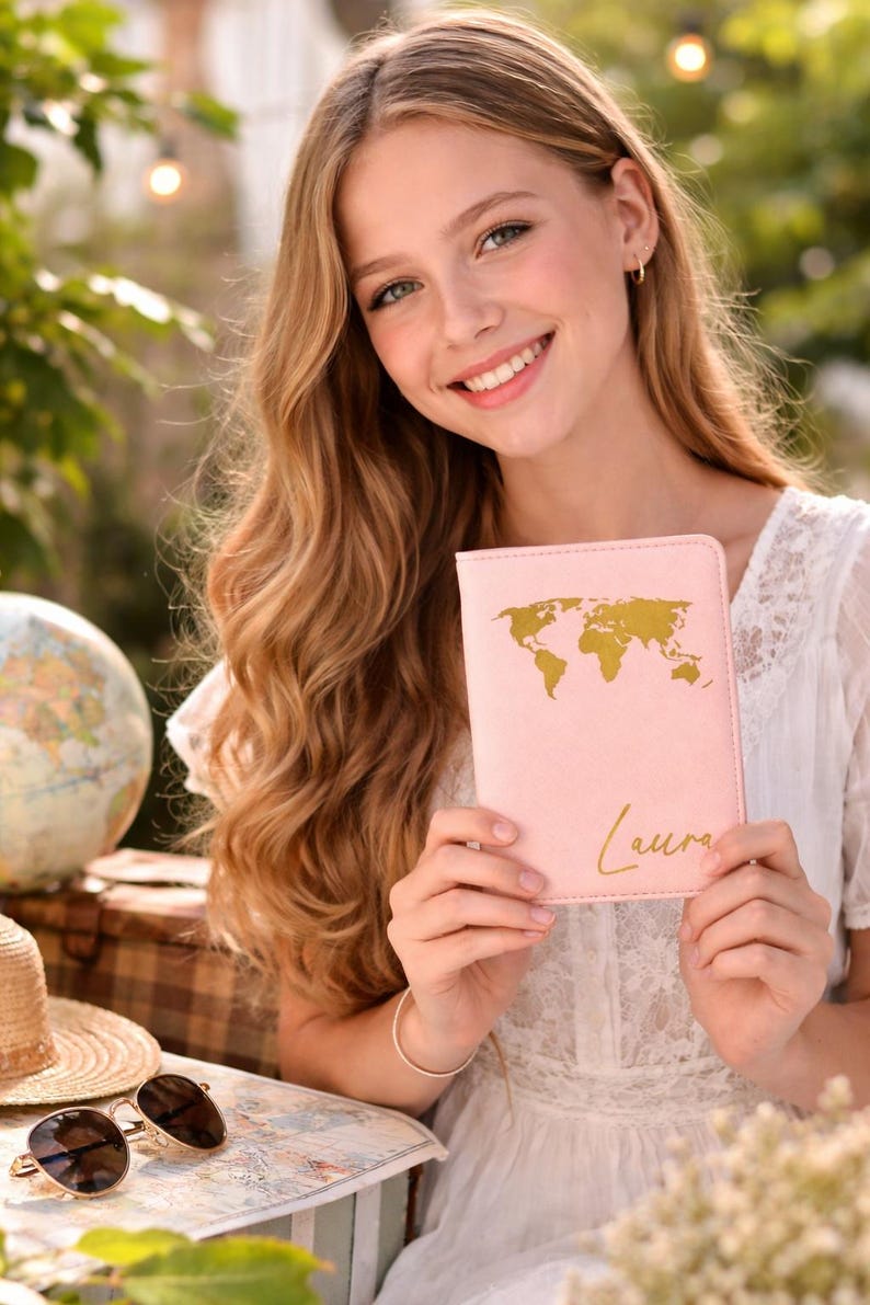 Glückliches Mädchen hält eine personalisierte rosa Reisepasshülle mit Weltkartenmotiv in den Händen vor modernem Reisehintergrund mit Globus und Accessoires