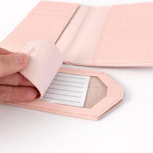 Detailaufnahme eines rosa Kofferanhängers mit geöffneter Schutzklappe und Sichtfenster sowie passender Reisepasshülle auf hellem neutralem Hintergrund