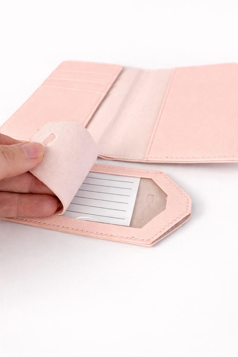 Detailaufnahme eines rosa Kofferanhängers mit geöffneter Schutzklappe und Sichtfenster sowie passender Reisepasshülle auf hellem neutralem Hintergrund