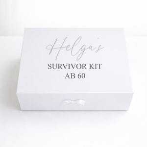 Weiße personalisierte Geschenkbox ‚Helga’s Survivor Kit ab 60‘ mit Schleife auf hellem, minimalistischem Hintergrund