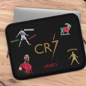 May include: Black laptop sleeve featuring images of a soccer player, the text "CR7 JAMES", and a goat illustration. The sleeve has a zipper closure and is designed to protect a laptop. The background is a wooden surface.