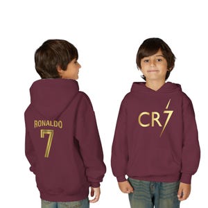 May include: Burgundy hoodie with gold text and graphics. The front features "CR7" with a lightning bolt, and the back displays "RONALDO" and the number "7". The hoodie has a hood and a front pocket.