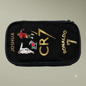 May include: A black rectangular pencil case featuring the gold text CR7, the name JOSHUA, and the name RONALDO with the number 7. The design includes two soccer player graphics in action poses and a small goat illustration in red, black, and green.
