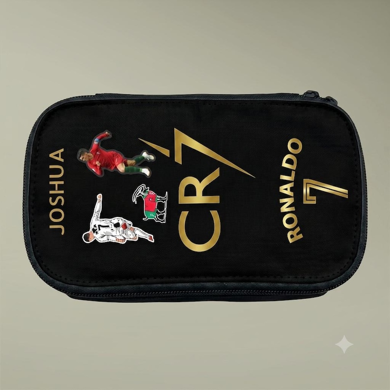 May include: A black rectangular pencil case featuring the gold text CR7, the name JOSHUA, and the name RONALDO with the number 7. The design includes two soccer player graphics in action poses and a small goat illustration in red, black, and green.