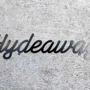 May include: Black metal sign with the word "Hydeaway" written in a cursive font.