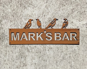 Könnte beinhalten: Ein rostiges braunes Metallschild mit dem Text "MARK'S BAR" in weißen Buchstaben. Vier schwarze Vogelsilhouetten sitzen oben auf dem Schild.
