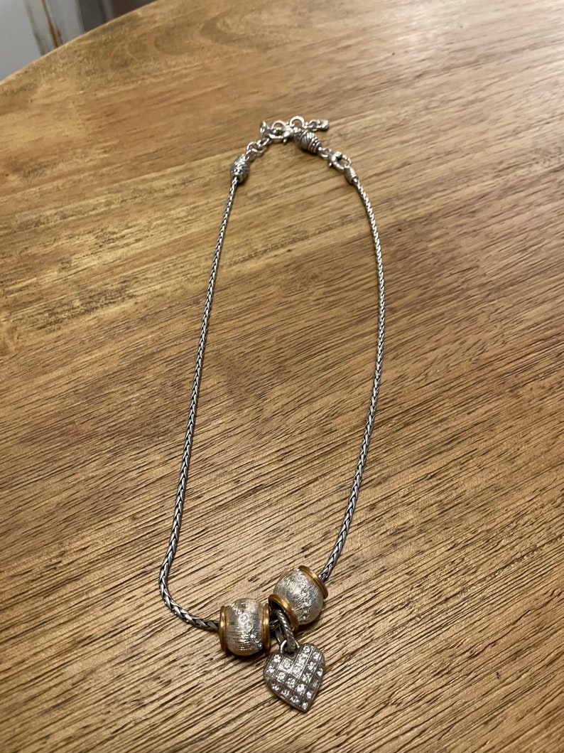 Pu&ograve; includere: Una collana a catena d'argento con tre perline d'argento e oro e un ciondolo a forma di cuore d'argento con strass trasparenti.