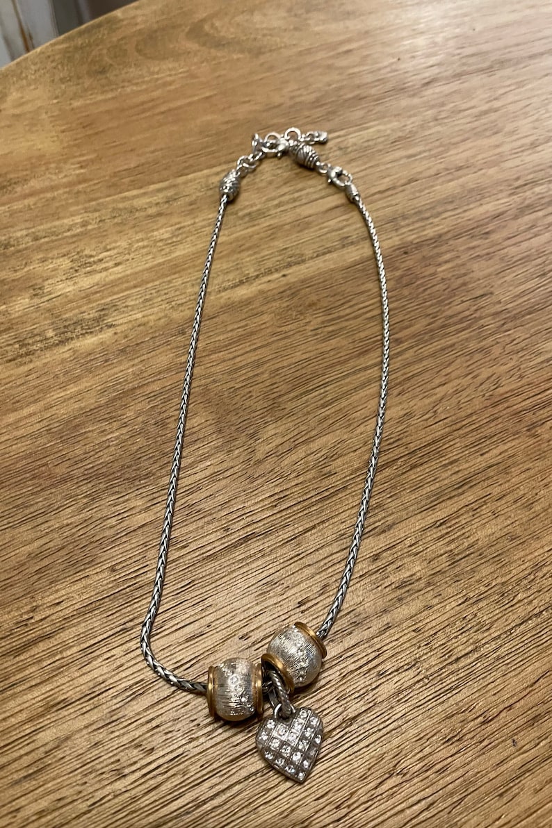 May include: A silver chain necklace with three silver and gold beads and a silver heart charm with clear rhinestones.
