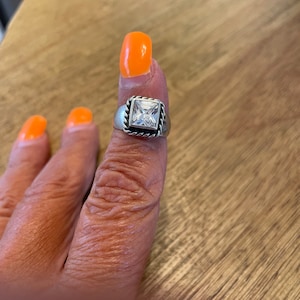 May include: A silver ring with a square-shaped clear stone set in a rope-like border.