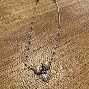Pu&ograve; includere: Una collana a catena d'argento con tre perline d'argento e oro e un ciondolo a forma di cuore d'argento con strass trasparenti.