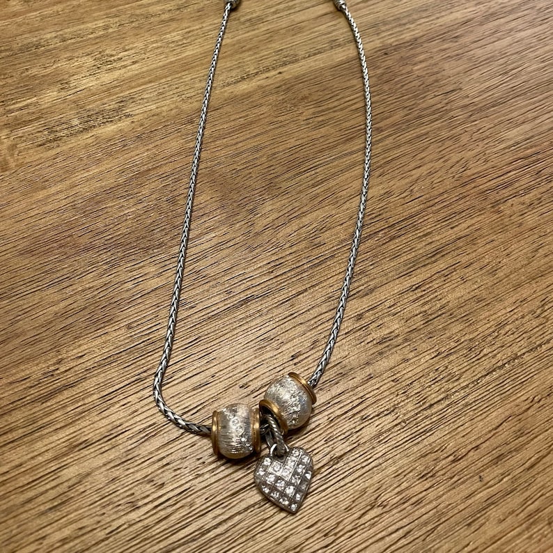Pu&ograve; includere: Una collana a catena d'argento con tre perline d'argento e oro e un ciondolo a forma di cuore d'argento con strass trasparenti.