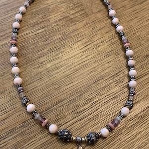 May include: A beaded necklace featuring a variety of pink, purple, and silver beads. The necklace has a pendant with a spiral design. The beads are strung on a silver chain and the necklace is displayed on a wooden surface.