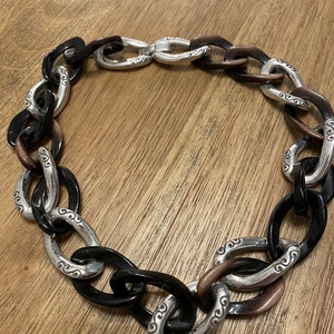 May include: A chunky silver and black chain necklace with a decorative design. The chain is made of large oval links with a silver metal center and black resin on the outside.