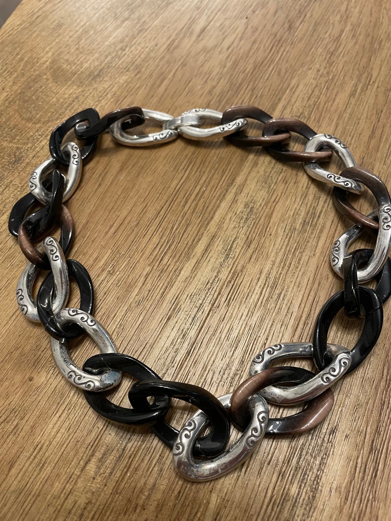 May include: A chunky silver and black chain necklace with a decorative design. The chain is made of large oval links with a silver metal center and black resin on the outside.
