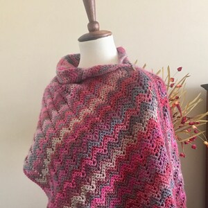 May include: A handmade, crocheted poncho in shades of pink, gray, and cream, displayed on a mannequin. The poncho features a chevron pattern and a cowl neck. The Carolina L. Ball signature is visible in the lower left corner.