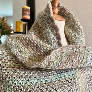 May include: A handmade, multi-colored crochet cowl scarf. The scarf is displayed on a mannequin, showcasing its intricate stitch pattern and blend of neutral tones, including gray, beige, and hints of blue and green. The cowl is designed to be worn around the neck.