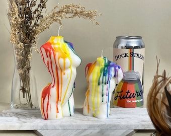 Op de afbeelding: Twee kleurrijke kaarsen in de vorm van menselijke torso's, met regenboog-druppelpatronen. De kaarsen staan op een wit marmeren oppervlak. Achter de kaarsen staat een blikje Dock Street Futuro Italiaans Pilsner bier.