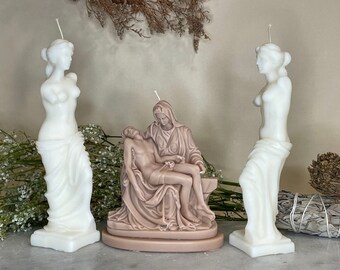 Op de afbeelding: Drie witte en bruine gebeeldhouwde kaarsen die de figuren van Venus de Milo en de Pieta voorstellen. De kaarsen zijn op een tafel gerangschikt met gedroogde bloemen en saliebundels.