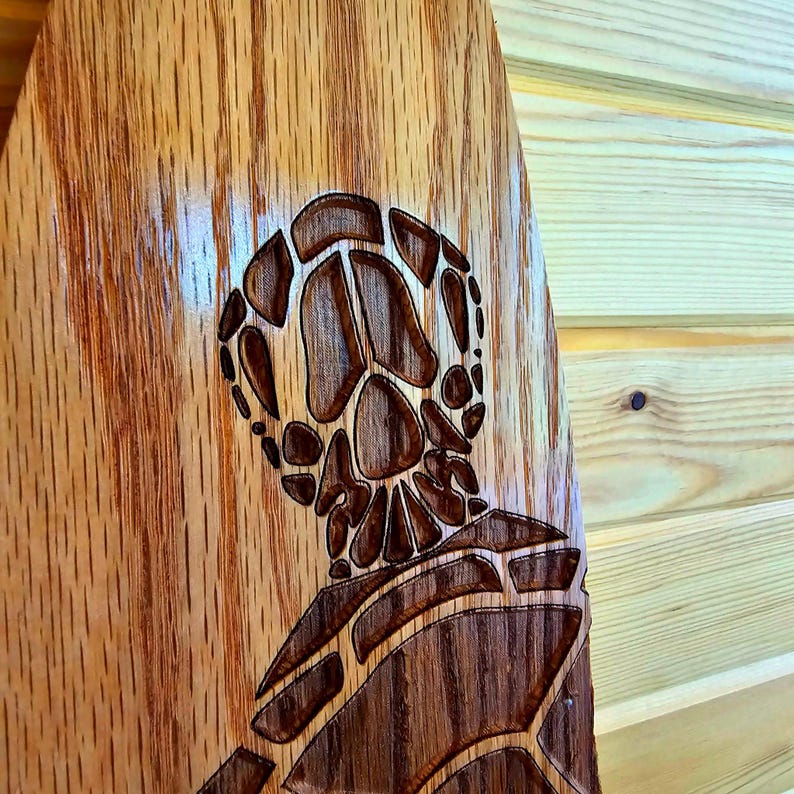 May include: A wooden paddle with a carved sea turtle design. The turtle is intricately detailed and has a brown and light brown color scheme.