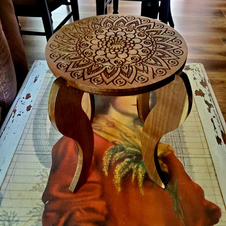 May include: A wooden stool with a round top featuring a laser-etched mandala design. The stool has three curved legs.