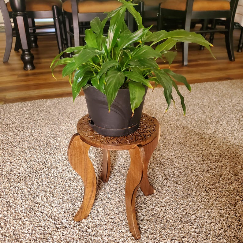 May include: A green peace lily plant in a black pot sits on a wooden plant stand with a carved mandala design. The stand has three legs and is on a light brown carpet.
