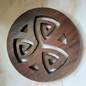May include: A round, wooden trivet with a carved Celtic knot design. The trivet is made of dark brown wood with a lighter-colored interior, creating a contrast. The design features a three-part knot with triangular cutouts around the edges.