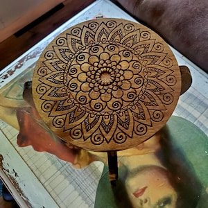 May include: A wooden stool with a round top featuring a carved mandala design. The mandala is intricate and detailed, with a brown wood finish.