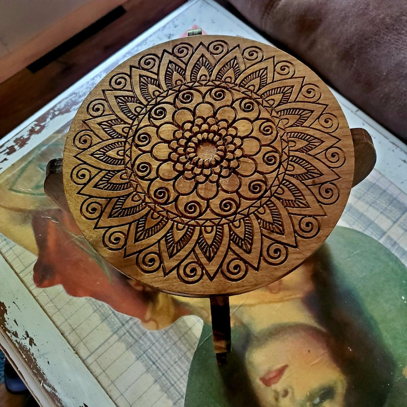May include: A wooden stool with a round top featuring a carved mandala design. The mandala is intricate and detailed, with a brown wood finish.
