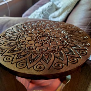 May include: A wooden stool with a round, carved top featuring a mandala design. The intricate pattern is carved into the wood, creating a detailed and decorative piece.