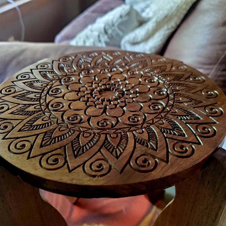 May include: A wooden stool with a round, carved top featuring a mandala design. The intricate pattern is carved into the wood, creating a detailed and decorative piece.