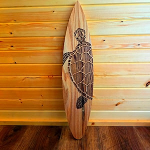 May include: A wooden surfboard with a carved sea turtle design. The surfboard is brown and has a natural wood grain.