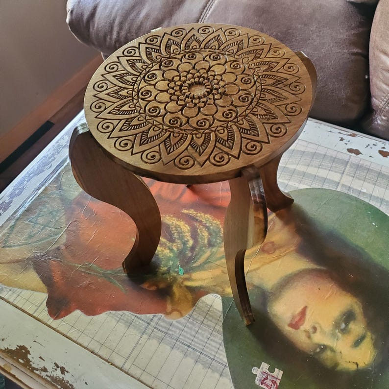 May include: A wooden stool with three legs and a round top. The top features a carved mandala design.