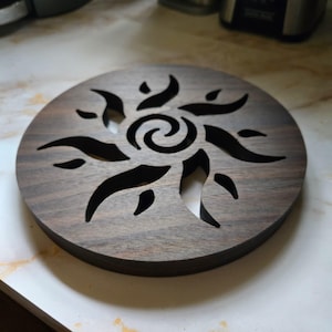 May include: A round, dark brown wooden trivet with a sunburst design cut out of the center. The trivet is made of a dark wood with a visible grain. The sunburst design features curved lines and a central spiral.