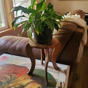 May include: A wooden plant stand with a carved top and three legs. A green plant in a black pot sits on the stand.