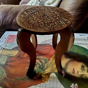 May include: A wooden stool with a round top featuring a carved mandala design. The stool has three legs with a curved shape.